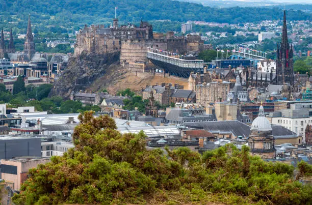 Cityscape of Scottish Capital city, in mid-summer,set up for Military Tattoo event.Coarse bushes at cliff edge. Landmark historic Edinbugh Castle,surrounded by iconic,landmarks St.Giles and The Hub.
