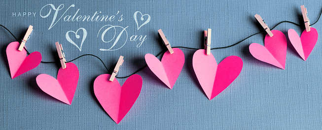 Pink paper cut hearts hanging on a clothesline with wooden clothespins. HAPPY VALENTINE'S DAY text is in the corner. Can be used as a design for Valentine's day holiday greeting cards or posters.