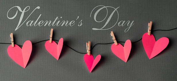 Pink paper cut hearts hanging on a clothesline with wooden clothespins. VALENTINE'S DAY text is in the center. Can be used as a design for Valentine's day holiday greeting cards or posters.