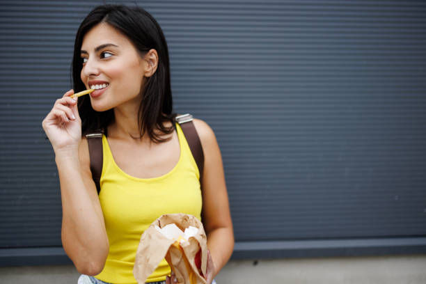 Woman eating fast food in the city Beautiful young woman leaning on wall and eating French fries while waiting in the city smiling potato stock pictures, royalty-free photos & images