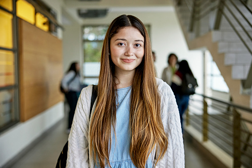 Waist-up view of longhaired woman in early 20s wearing casual attire, standing in hallway, and looking at camera with shy smile. Property release attached.