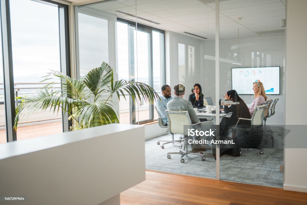 Multiracial Start Up Team Making Plans Wide angle shot of a multiracial team of men and women sitting in a closed meeting room of a modern office. Shot through glass walls, big potted plant next to large windows onto the patio. Office Stock Photo