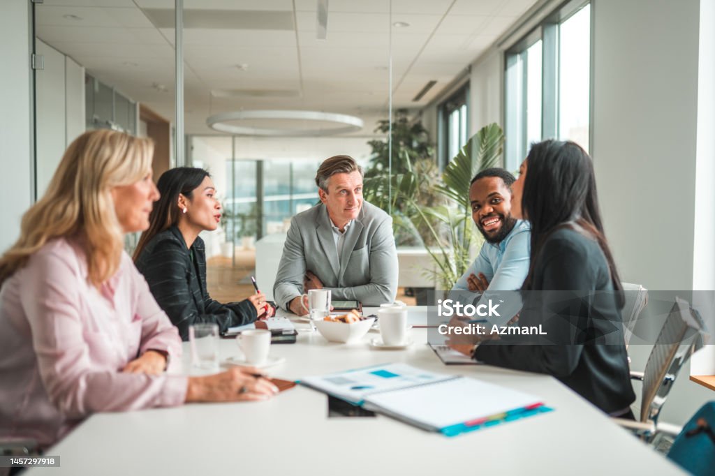 Successful Mid Adult CEOs Meeting in International Corporation Multi-ethnic team of mid adult male and female managers sitting in a meeting room, leaning on the table and discussing. Waist up image, front view of the Caucasian and African man, side view of the Caucasian and Asian women. Meeting Stock Photo