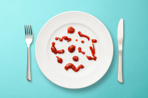 Overhead shot of white plate with spilled ketchup and silverware on light blue background.