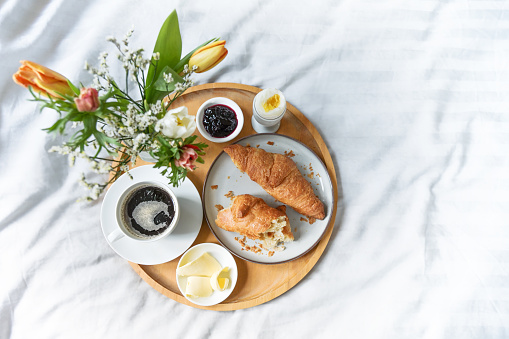 Lazy breakfast in bed, wooden tray with croissant, coffee, jam, egg, butter and flowers on gray green white bed linen, Sunday or holiday morning, copy space, top view form above, selected focus, very narrow depth of field