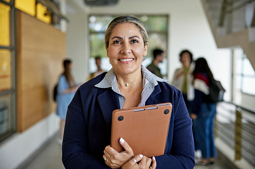 Waist-up view of late 40s woman standing in university hallway, holding laptop, and smiling at camera. Property release attached.