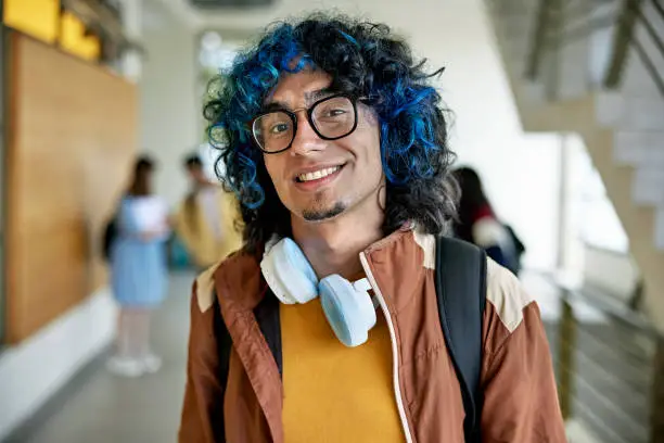 Early 20s Hispanic man with blue dyed hair, eyeglasses, and headphones around his neck pausing in hallway to grin at camera. Property release attached.