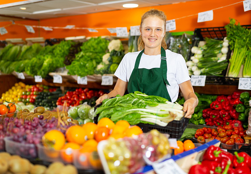 Smiling teenage girl in uniform working in grocery shop as job experience, posing with salad