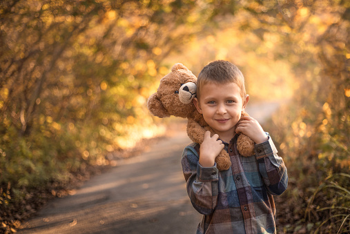 a boy stands on a country road in the forest on his shoulders sits a toy bear