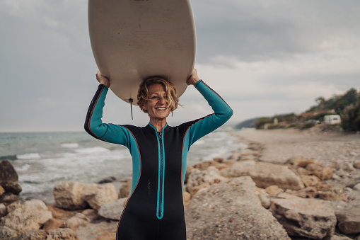 Portrait of a mature female surfer in wetsuit, holding a surfboard on her head on the beach.