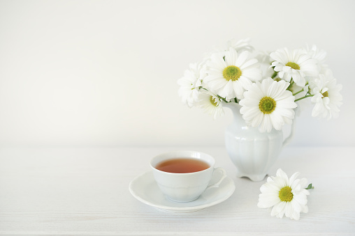 Bouquet of white flowers and cup with tea on white wooden table, copy space. Herbal tea and bouquet of daisies or chamomile flowers on a white background
