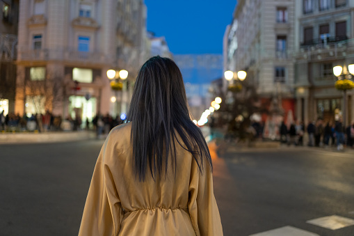 Back view of unrecognizable female with long dark hair in beige coat standing on asphalt crowded street illuminated with lanterns near old buildings at night