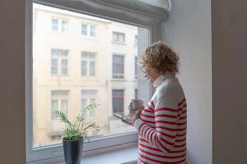 Adult woman drinking coffee and looking at her smart phone in front of the window of her house.