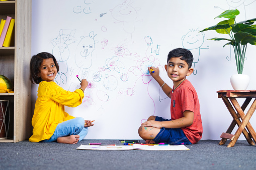 Young siblings kids drawing with crayon color on wall by looking at camera at home - concept of mischief, troublesome children and childhood lifestyles