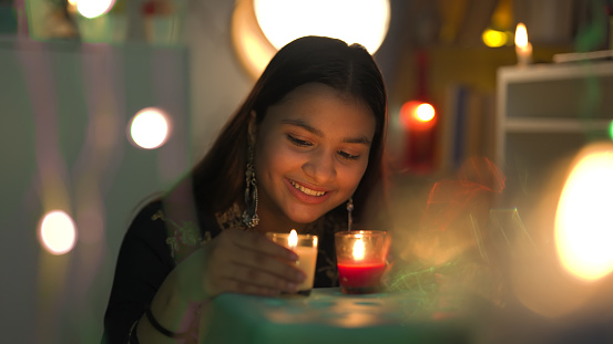 Smiling pretty teenage wearing big earrings sitting and hold burning glass candles in hand closer to face on the occasion of Diwali. woman enjoy Deepawali festival with Decorative surroundings