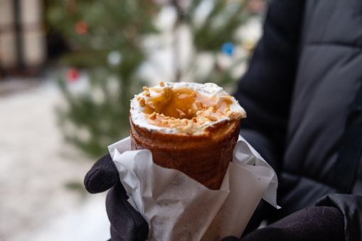 A man holds a trdelnik with white cream and caramel in his hands, selective focus.