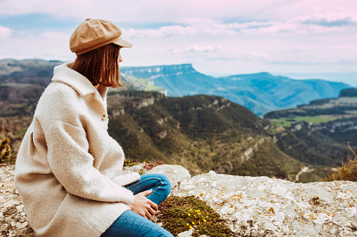 Rear view portrait of a young stylish woman wears beige beret and coat, sitting on the mountain. Active traveler lifestyle climbing mountain peak, enjoying the beautiful landscape.