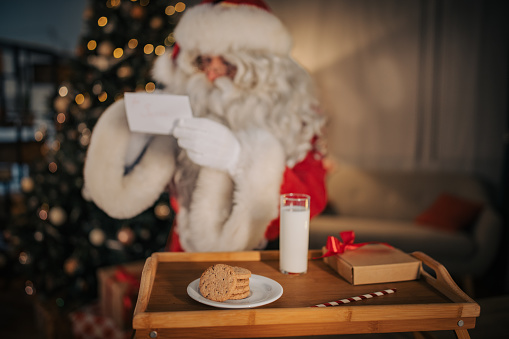 Santa Claus reading a note, cookies and glass of milk that is left for him on a tray at home.