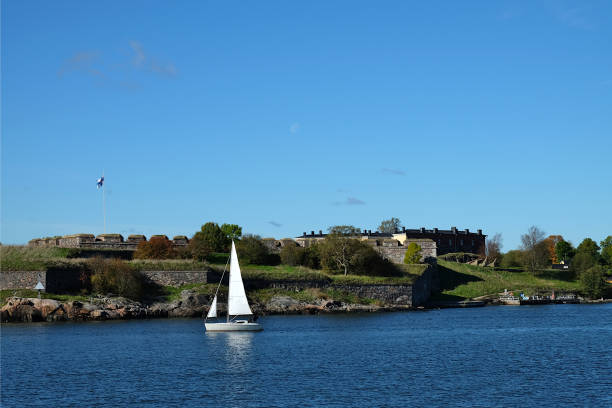 View over the strait between two islands with autumn colored trees behind and moving sail boat, Vallisaari, Helsinki Biennial 2021