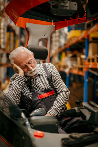Exhausted storage male worker sleeping during working hours. Senior forklift driver napping in a forklift