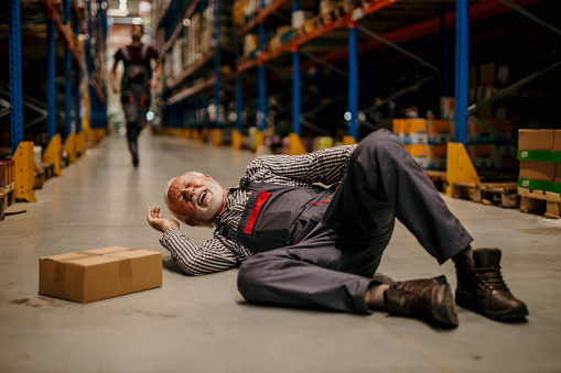 A warehouse worker had an accident in the storage. A man in uniform lying down on the ground while his coworker is in the background running toward him.
