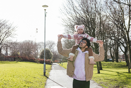 A waist up medium front view of a father and his adopted daughter making memories in a public park in the North East of England. She is smiling and sitting on her fathers shoulders with her arms outstretched as they walk through the park.
