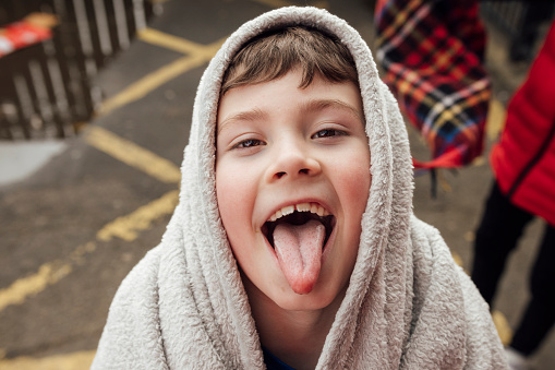 A point of view of a young boy who is looking into the camera and sticking his tongue out towards the camera. He has a blanket on his head to keep him warm in thge cold weather in the park in the North East of England.
