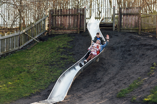 A wide-angle front view of a father and his adopted children enjoying a slide in a public park in the North East of England. There are three of them sliding down together with big smiles on their faces.