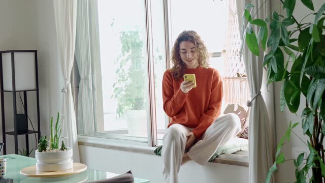 Happy young woman using cell phone sitting on windowsill at home.