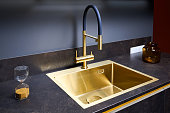 Luxurious interior square golden brass sink and faucet double tap mixer in contemporary modern design with stone marble stoneware countertop black and gold kitchen with sandwatch and glass pot.