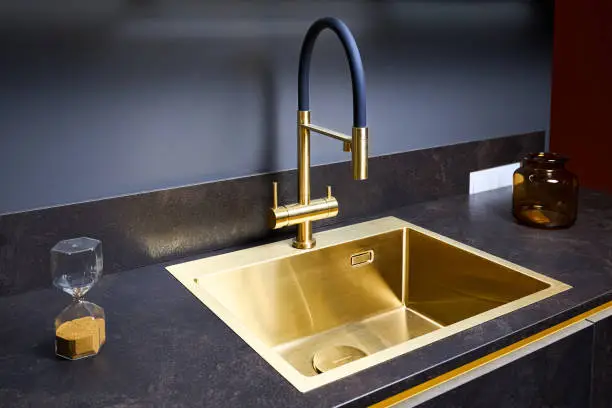 Luxurious interior square golden brass sink and faucet double tap mixer in contemporary modern design with stone marble stoneware countertop black and gold kitchen, with sandwatch and glass pot.