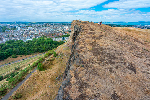 Edinburgh,Scotland-August 01 2022: Visitors climbing to Arthur's seat,stand at the precipice of a high vertical cliff in awe of views across Edinburgh City,on a summer day in Scotland's capital.