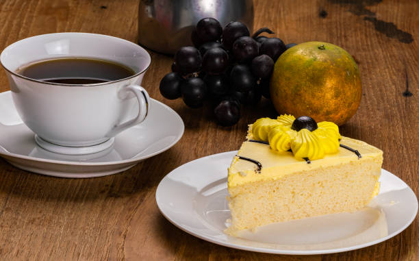 Closeup view slice of delicious homemade sponge cake in white ceramic dish with a cup of black coffee in white ceramic cup, bunch of grapes, an orange and moka pot coffee maker on wooden table.