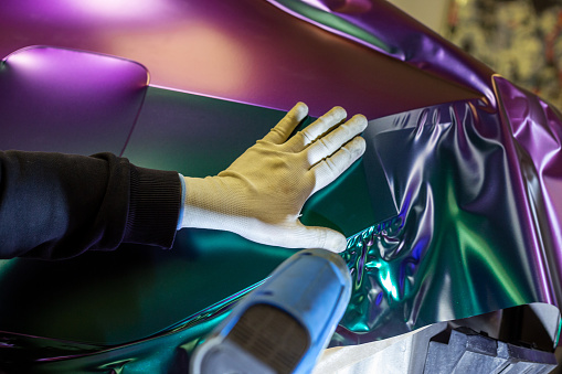 The process of wrapping a car with chameleon-colored vinyl film. A car wrapping specialist covers the car body with vinyl sheet or film. Selective focus.