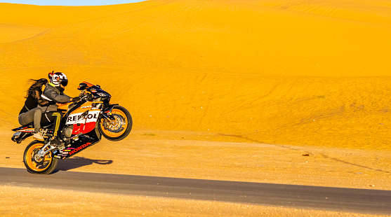 Saoura, Algeria - December 28, 2022: a biker rearing up his Repsol Honda motorcycle with a young girl with no helmet holding him on the back in a desert road in Taghit, Bechar in Algeria. Sunny day with sand dune and blue sky.