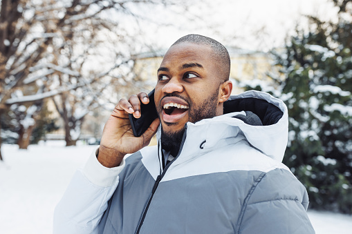 Happy surprised African American man in casual white sport clothes talking on phone, smiling and shocked in park or street outdoor. Winter season with snow