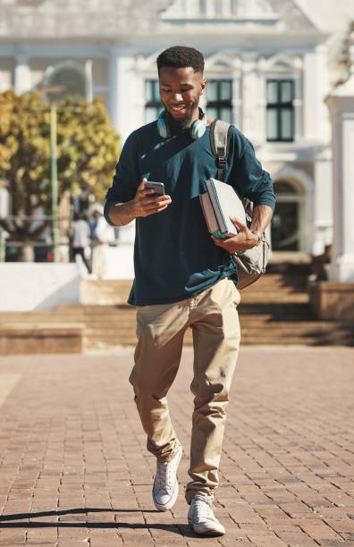 university, education and phone with a black man student typing a text message while carrying books. contact, study and scholarship with a male pupil walking to class at college for development - adult education full length book imagens e fotografias de stock