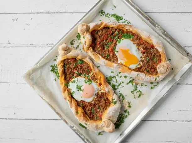 Delicious filled turkish pide or flatbread with spicy vegetable, minced meat filling. Topped with a oven poached egg. Served on a baking tray isolated on white background. Flat lay
