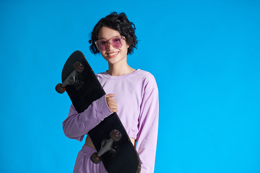 Portrait of smiling teenage girl holding skateboard, isolated on blue