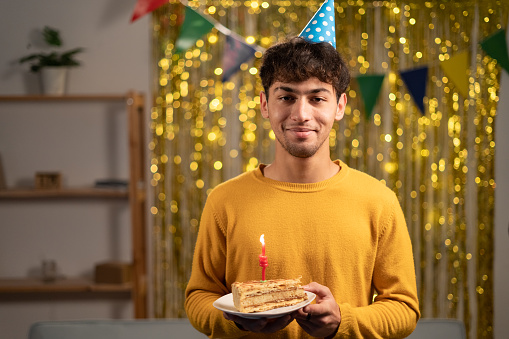 Young handsome man holding birthday sweet cake over golden home background with a happy face standing and smiling with a confident smile. Copy space