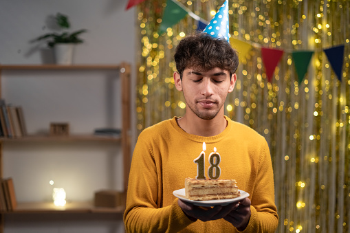 Young man in cone party hat to make wish holding his birthday cake with candles number 18 during celebration at home. Eighteens birthday concept