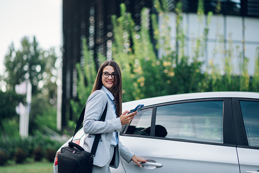 Business portrait of young professional businesswoman in suit standing outside office building and going inside car while using smartphone. Successful female going to a meeting by car. Copy space.