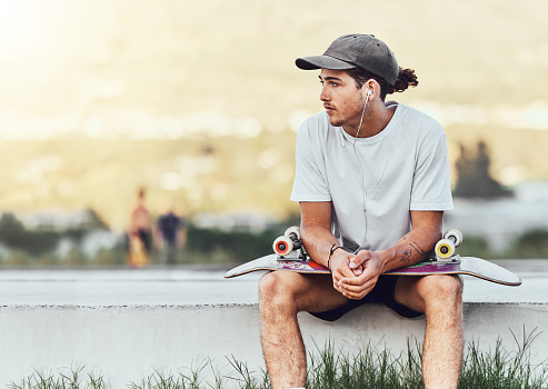 Young man, skateboard and relax outdoor, thinking or chill being casual, trendy and earphones. Male, skater or idea for break, calm and cool look being edgy, content and hipster with style on weekend