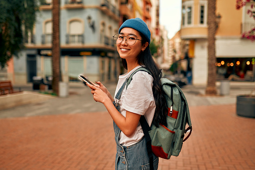Asian young woman student in glasses with a backpack and a hipster hat goes to study at the university using a smartphone. Woman tourist walking through the streets of the city.