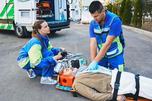 EMS, first aid. Two paramedics performing closed-chest cardiac massage for victim man who lies unconscious on medical stretcher near ambulance outdoor