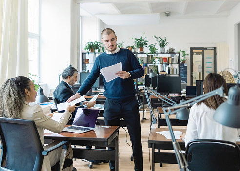 Man giving handouts to diverse employees in open plan office. Male employee giving some documents to a woman sitting at her desk in open plan office.