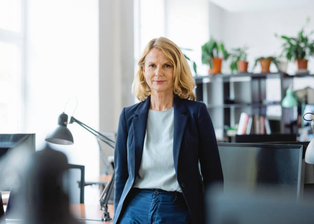 portrait of a confident mature businesswoman standing by a desk in office - leadership business women senior adult imagens e fotografias de stock