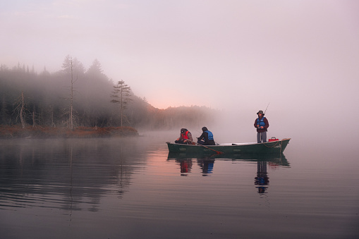 3 men fishing in a canoe very early in the morning. A very thick fog covers the lake revealing the sunrise. One person fishes standing up, while the others set up their fishing rods.