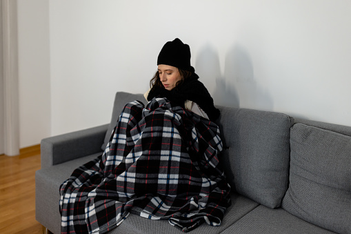 A young Caucasian woman with heating problems sitting on sofa wrapped in plaid and wearing a hat