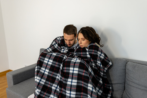 Two poor people sitting together on sofa wrapped in plaid blanket trying to warm up